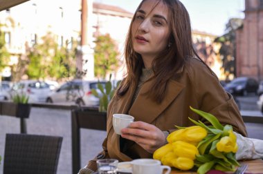 Genç ve güzel bir kız caddedeki bir kafede espresso içiyor. Çerçevede bahar güneşi. Masanın üzerinde bir buket çiçek yatıyor. Bir fincan kahve için sabah molası. Uzayı kopyala yatay olarak resim