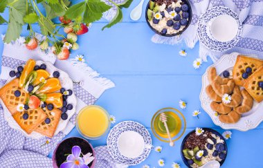 Yukarıdan çekilmiş Paskalya kahvaltısı sahnesi. Çay fincanları, reçelli pasta, kurabiyeler, taze portakal suyu, meyveler, böğürtlenli granola, balım, masada yapraklı çilek ve meyveli..