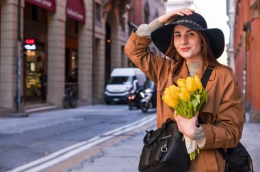 Sarı lalelerle dolu buketi olan güzel genç bir kadın. Şapkalı, trençkotlu bir kız çiçeklerle İtalya 'nın eski şehrinde yürüyor. Turizm ve eğlence kavramı.