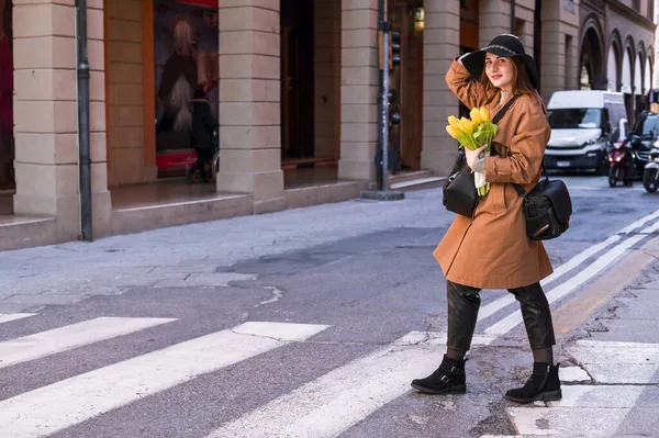 Belle jeune femme avec un bouquet de tulipes jaunes. Fille coiffée d'un chapeau, une tranchée traverse la route de la ville d'Italie avec des fleurs à la main. Le concept de tourisme et de divertissement. — Photo