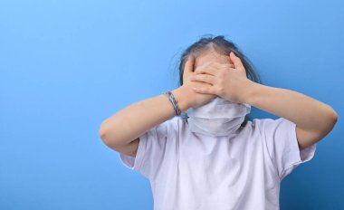 Koronavirüslü bebek. Dünya salgını konsepti COVID 19. Koruyucu tıbbi maskeli küçük bir kız elleriyle gözlerini kapatıyor. Fotoğraftaki çocuğun duygularına. Boşluk ve blu arkaplanı kopyala 