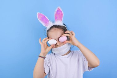 Coronavirus ve Paskalya tatili. Koruyucu tıbbi maskeli ve dekoratif tavşan kulaklı bir çocuk Paskalya yumurtalarını elinde tutuyor. Küçük kız ve Paskalya sembolü. Dünya salgını. Boşluğu kopyala