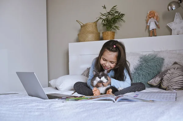 Distance learning online education. Schoolgirl with digital tablet laptop notebook and doing school homework. Sitting on bed with training books. Home school. A child with a pet. Copy space