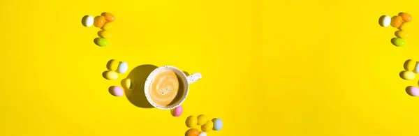 A lot of multi-colored Easter eggs and a mug of coffee with froth on a yellow background. Happy Easter card with copy space for text. Minimal style. In a hard light. selective focus. Above. Banner.