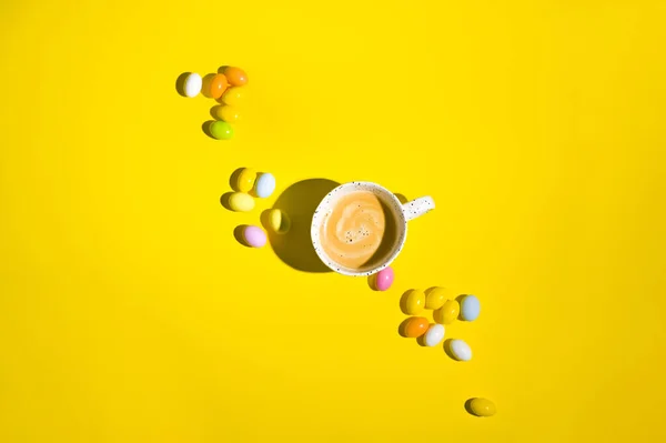 A lot of multi-colored Easter eggs and a mug of coffee with froth on a yellow background. Happy Easter card with copy space for text. Minimal style. In a hard light. selective focus. Art style
