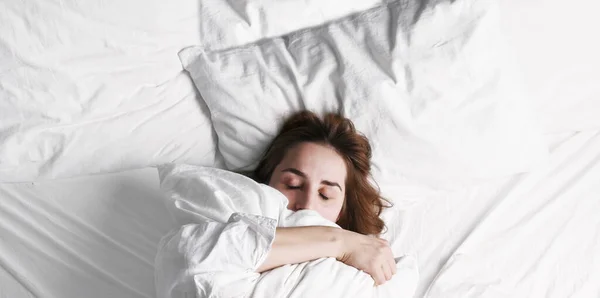 Girl wakes up in the morning. A young woman in bed hugs a pillow snugly and comfortably. Shows no desire to wake up. White bed, blanket and pillows. Free space for text. Copy space. Banner