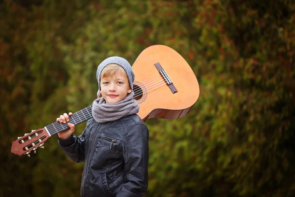 Симпатичный маленький мальчик с гитарой на природе. Он одет. — стоковое фото