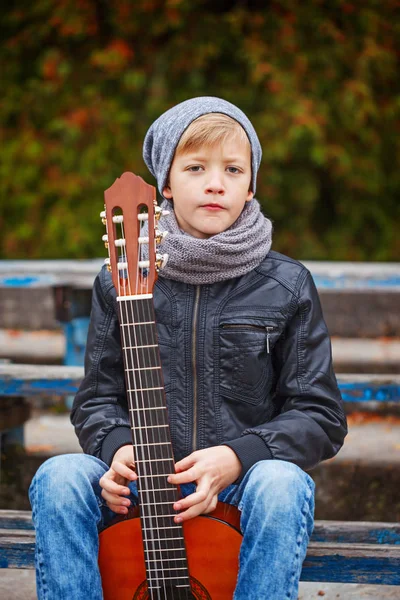 Cute little boy with guitar in autumn day. He is dressed in a bl — Stock Photo, Image