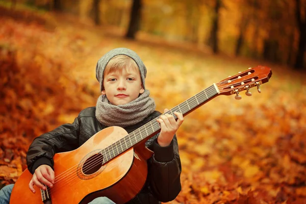Liten pojke som spelar gitarr på natur bakgrund, höstdag. Barnens intresse för musik . — Stockfoto