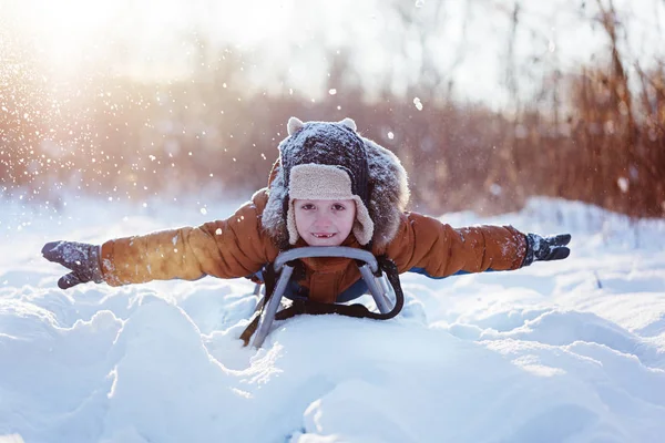 Menino engraçado feliz em roupas quentes de inverno se divertindo no trenó de neve, ao ar livre durante a queda de neve — Fotografia de Stock