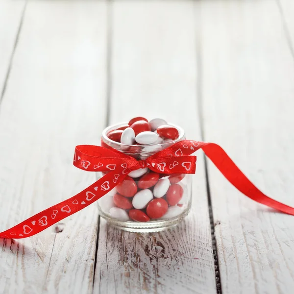 Colorato barattolo di caramelle decorato con un cuore di fiocco rosso su sfondo di legno bianco. Concetto di San Valentino — Foto Stock