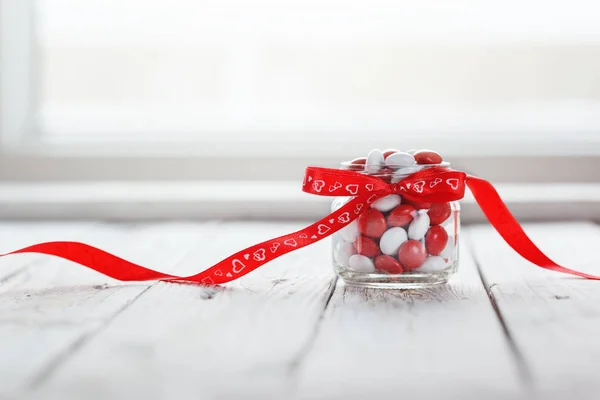 Colorato barattolo di caramelle decorato con un cuore di fiocco rosso su sfondo di legno bianco. Concetto di San Valentino — Foto Stock