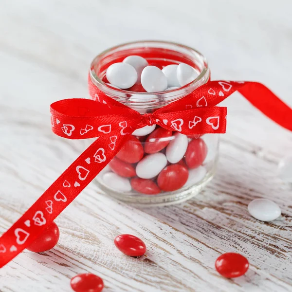 Colorful candy jar decorated with a red bow  hearts on white wooden background. Valentines day concept — Stock Photo, Image