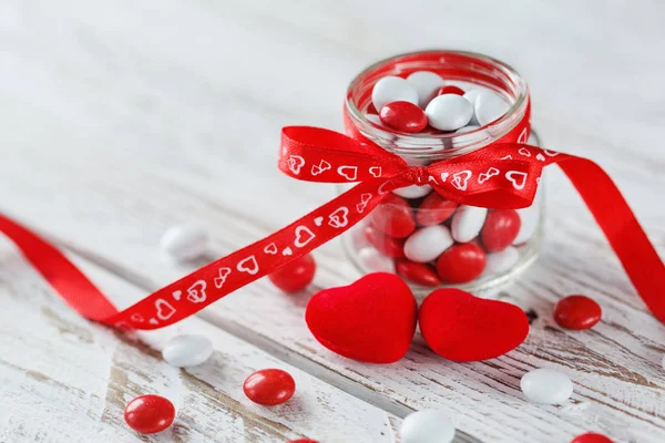 Colorful candy jar decorated with a red bow  hearts on white wooden background. Valentines day concept — Stock Photo, Image