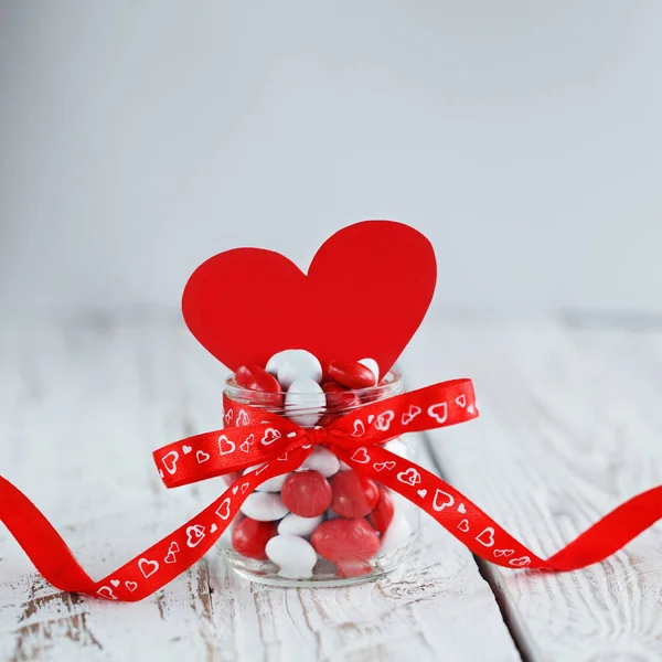 カラフルなキャンデーの瓶は赤いリボンと白い木製の背景に赤の紙のハートで飾られて。バレンタインの日の概念 — ストック写真