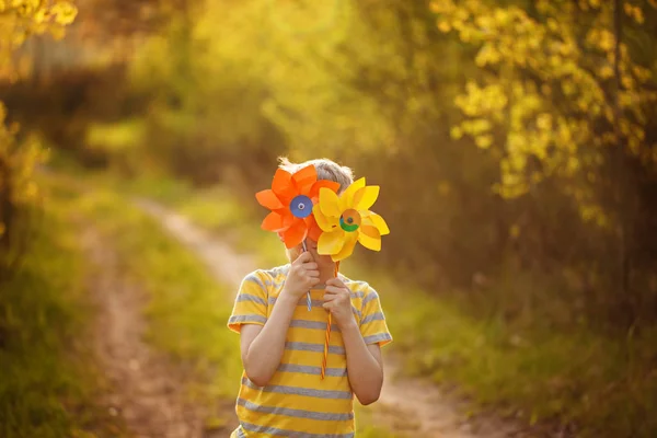 El niño se esconde detrás de molinos amarillos y anaranjados sobre el fondo verde del bosque en un día soleado . — Foto de Stock