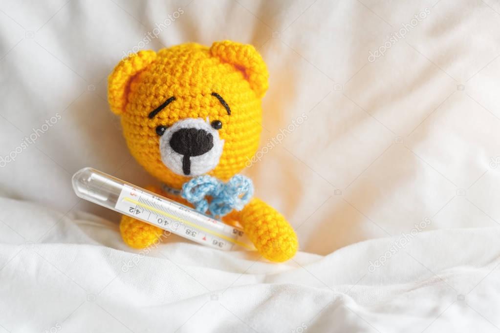 Yellow ill teddy bear with thermometer in white bedroom.