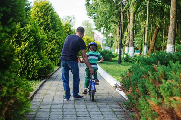 Ojciec pomóc synowi jeździć rower w lato park. Widok z tyłu — Zdjęcie stockowe