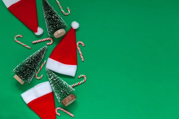 Noel kompozisyonu. Noel ağaçları ve Noel Baba şapkaları ve yeşil arka planda şekerler. — Stok fotoğraf