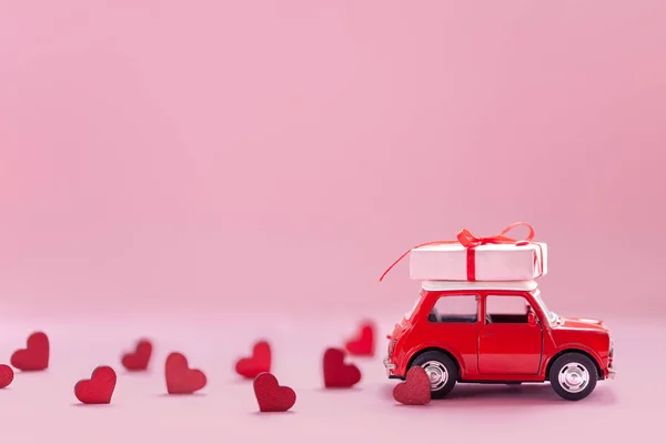 Красная игрушечная машина доставляет на крыше подарочную коробку с маленькими красными сердечками на розовом фоне. День святого Валентина . — стоковое фото