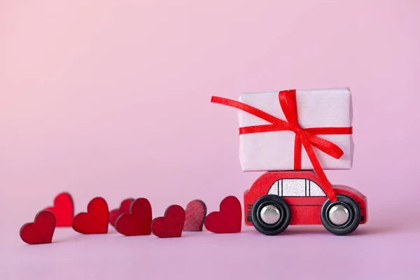 День святого Валентина фон. Миниатюрный красный автомобиль с коробкой подарков на крыше с красными сердцами на розовом фоне — стоковое фото