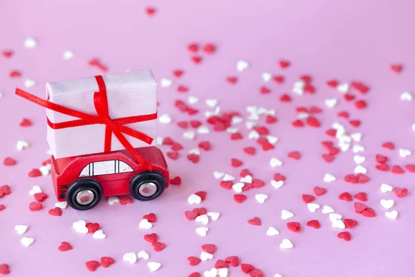 Миниатюрный красный автомобиль с коробкой подарков на крыше на розовом фоне с красными и червями — стоковое фото
