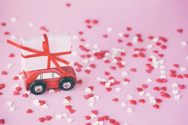 Миниатюрный красный автомобиль с коробкой подарков на крыше на розовом фоне с красными и червями wihte. Тонирование изображения — стоковое фото
