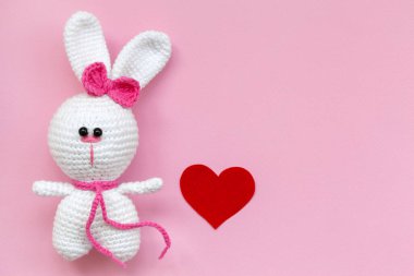 Pembe arka planda kırmızı kağıt kalbi olan küçük bir oyuncak tavşan. Düz yatış, üst görünüm.