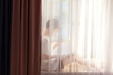 Evdeki pencerenin üzerinde oturan sevimli küçük çocuk kitap okuyor. Uzaklık öğrenimi çevrimiçi eğitim