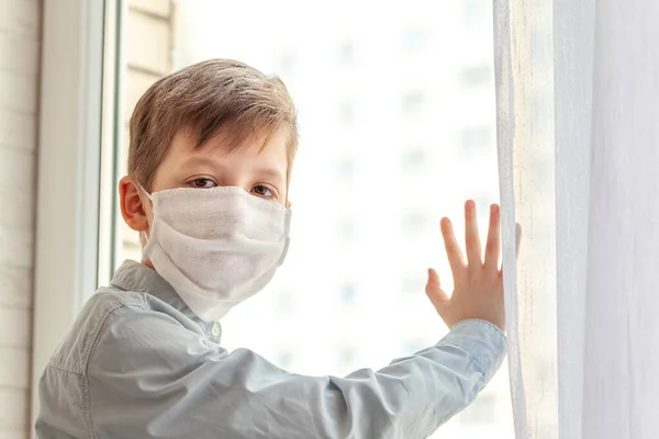 Child in a medical mask looking through the glass to the street. World coronavirus pandemic, social isolation, quarantine concept