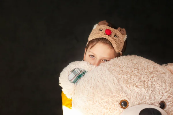Little sad child in a sleep mask hugs a toy of a big teddy bear. Sleep time concept.