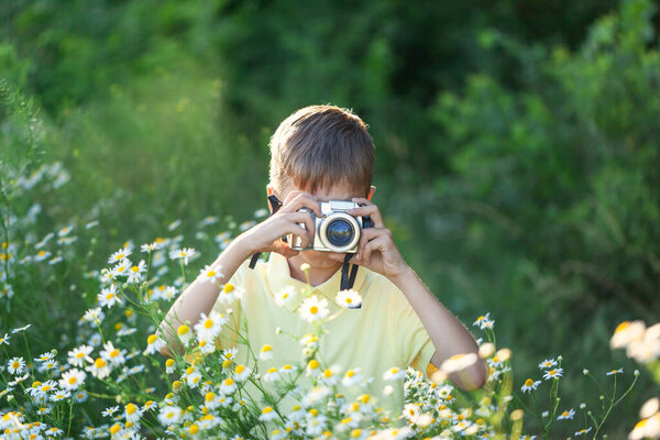 Child with professional camera is shooting flowers in sunny summer day on nature. Kid takes a photo in the camomile flowers field.