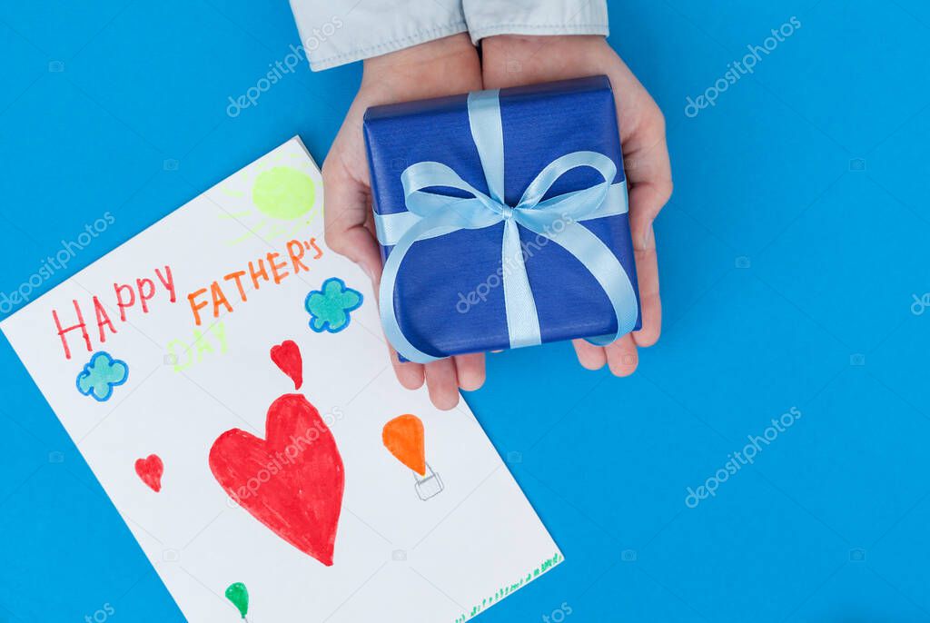 Kid's hands holding gift box with child's drawing on blue background. Concept Father's Day greeting card.