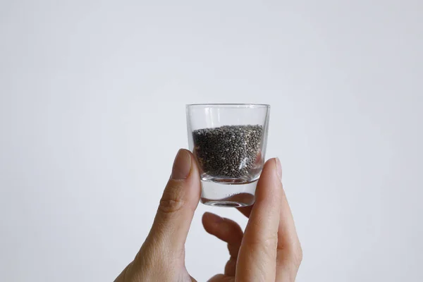 Beyaz arka planda gün ışığında bardakta chia tohumları olan bir kadın eli.
