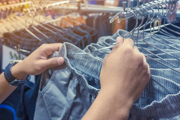 La gente controlla e sceglie pantaloni da acquistare su appendini in un negozio di abbigliamento. Focus selettivo sulla mano destra . — Foto Stock
