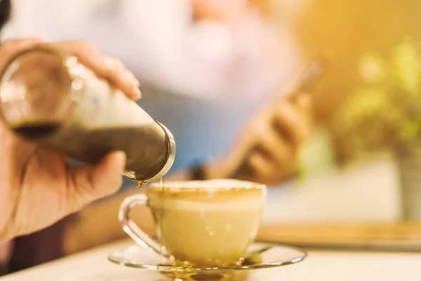 한 여성이 몰래 스마트폰을 하는 동안 커피 잔의 냄비에 시럽을 부었다. 선택적으로 병의 가장자리에 초점을 맞추고 — 스톡 사진