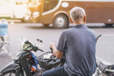Yaşlı bir adamın yol kenarına park etmiş bir motosikletin üzerinde oturması ve haberleri izlemek için telefon kullanması..