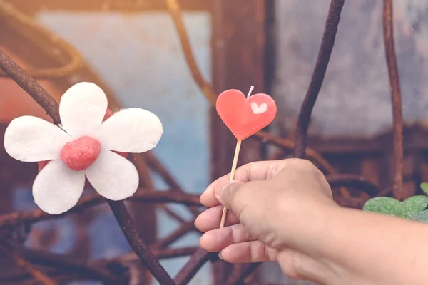 Eine rote herzförmige Kerze auf der Hand der Frau und eine künstliche Blume aus weißen und roten Kerzen mit einem bläulichen Bild rostiger Stahlstangen im Hintergrund. — Stockfoto