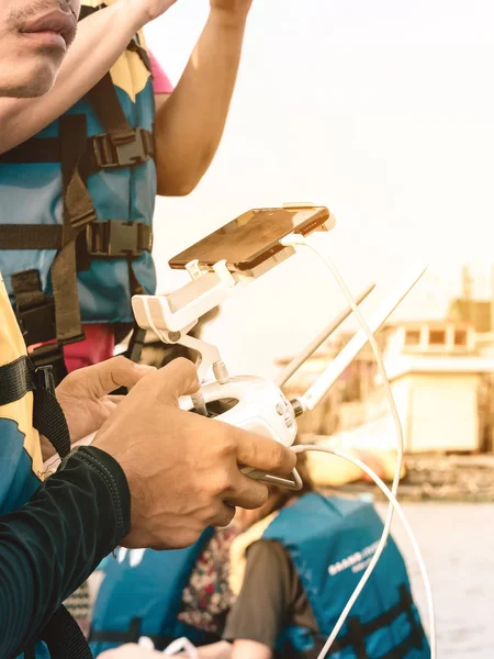 Joven con chaleco salvavidas azul y amarillo controlando un dron para tomar fotos de la costa del océano con los turistas mientras viaja en una balsa en el mar. Manos sosteniendo el mando a distancia del dron . — Foto de Stock