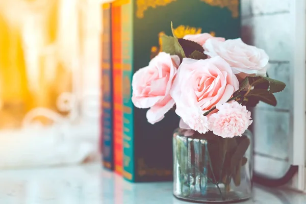 Belas rosas rosa artificiais em vaso de vidro em uma mesa branca com imagem azulada de três livros no fundo . — Fotografia de Stock