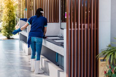 Kadın hademe kumaş ve deterjan kullanıyor. Erkekler tuvaletinin önünde lavabo ve musluk açıyorlar..