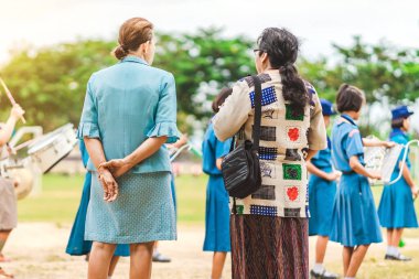 Asyalı bayan öğretmen, sabah okuldaki ulusal bayrağa saygılarını sunmak için sırada beklerken müzik çalan öğrencileri kontrol ediyor..