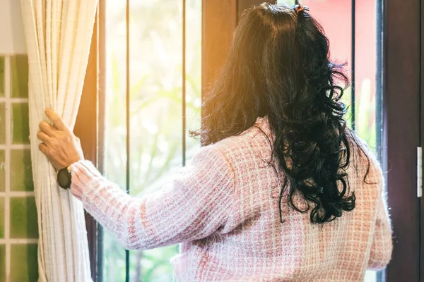 Back view of woman wear pink cardigan opens the window curtains and looks at the outside while at home to stop the spreading of the corona virus (covid-19).