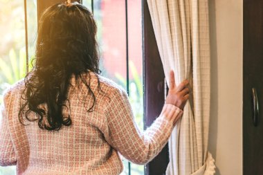 Pembe hırka giyen bir kadının arka görünüşü pencere perdelerini açar ve Corona virüsünün yayılmasını önlemek için evde iken dışarıya bakar (covid-19).