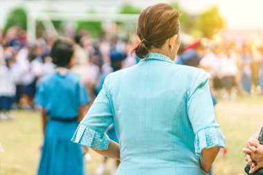 Asyalı bayan öğretmen, sabah okuldaki ulusal bayrağa saygılarını sunmak için sırada beklerken müzik çalan öğrencileri kontrol ediyor..