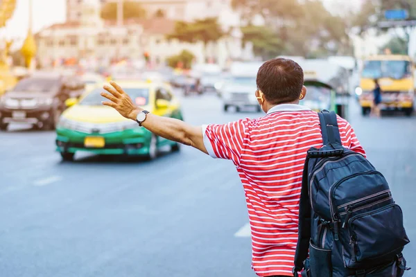 Back view of male patient with mask in red and white shirt with a black backpack standing at bus stop and waving his hand for Taxi or Bus in the city to go to the hospital.