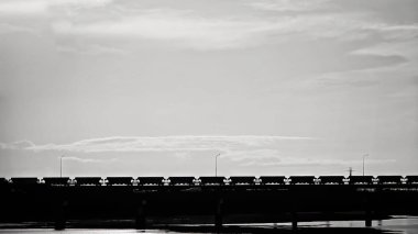 Hedefler arasında kargo taşıyan bir tren köprünün yanındaki bir nehri geçerken çarpıcı bir şekilde siyah ve beyaz renkte çizilmiş.