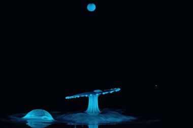 İki mavi su damlası çarpışır ve bir şemsiye şekli oluşturur ve yukarıdan gelen bir diğeri yüzeye baloncuk olarak iner, siyah arkaplan