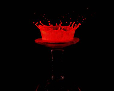 A red drop of milk splashed onto an upturned miniature wine glass creates a crown shaped splat against a black background clipart