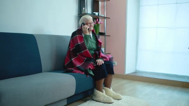 Abuela hablando por teléfono sentada en un sofá envuelta en un cuadros a cuadros — Vídeo de stock